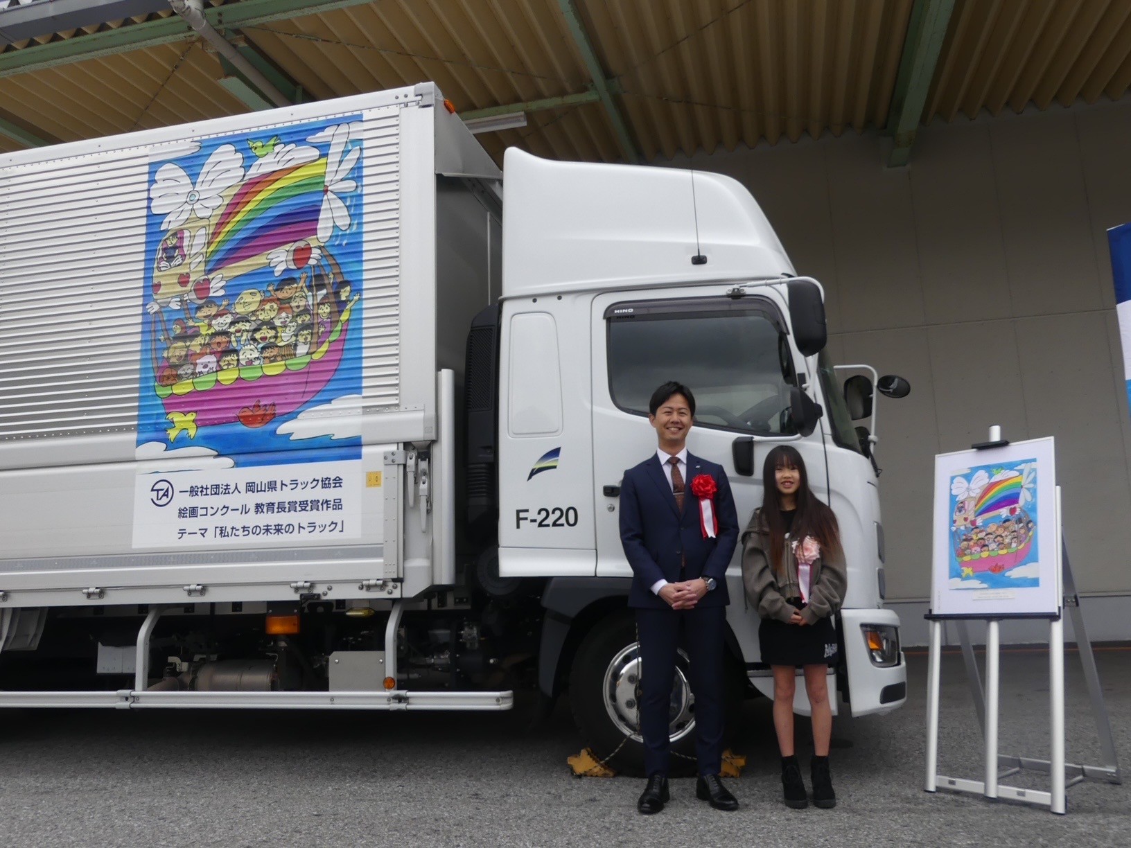 ラッピングトラックをバックに藤森運輸の藤森専務㊧と最優秀賞受賞者の真壁さん