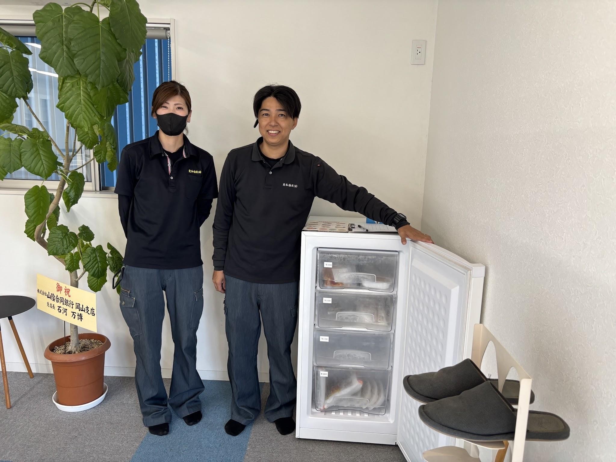 社食サービスの冷蔵庫の横に立つ健康経営担当の中野氏㊧と配車担当の西田氏